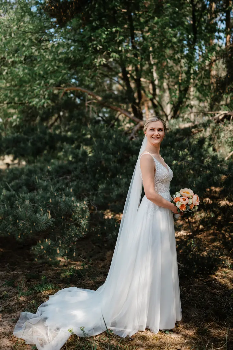 Braut im Brautkleid mit langer Schleppe im Halbschatten des Waldes