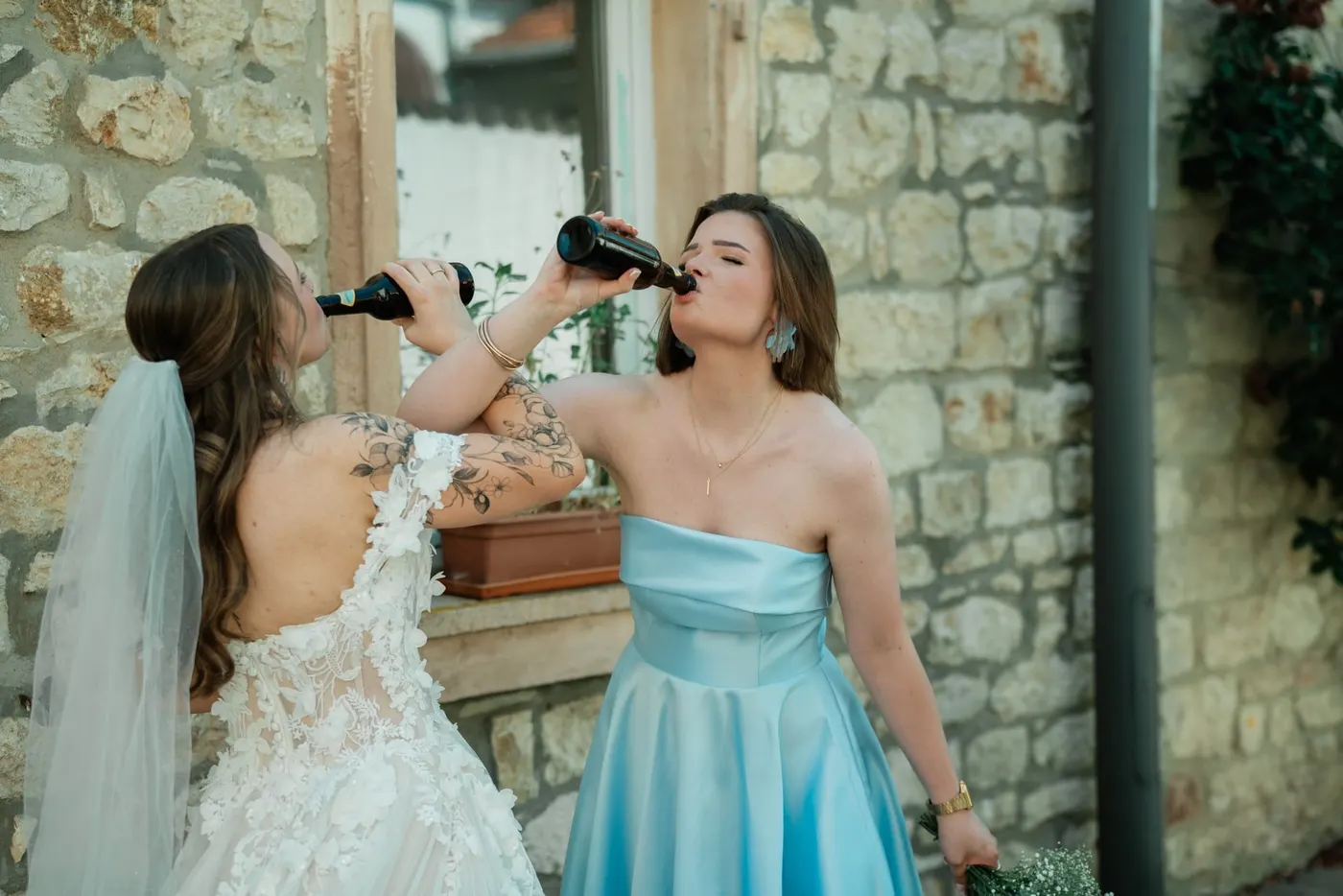 Braut und Trauzeugin trinken lachend aus Sektflaschen vor einer Sandsteinmauer