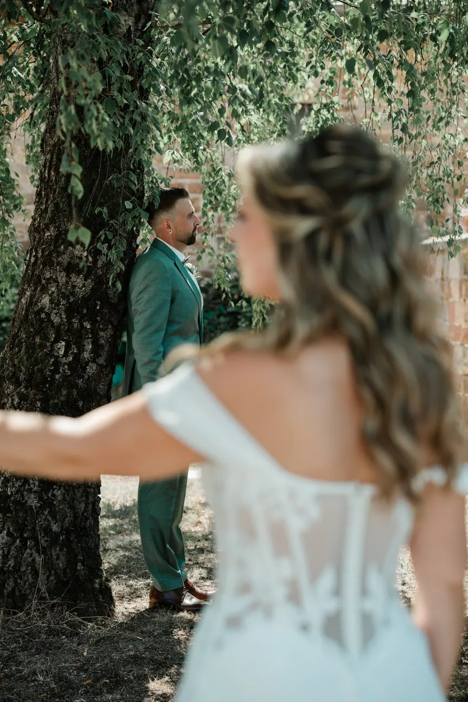 Braut mit offenem Haar blickt zu ihrem Bräutigam, der an einem Baum lehnt