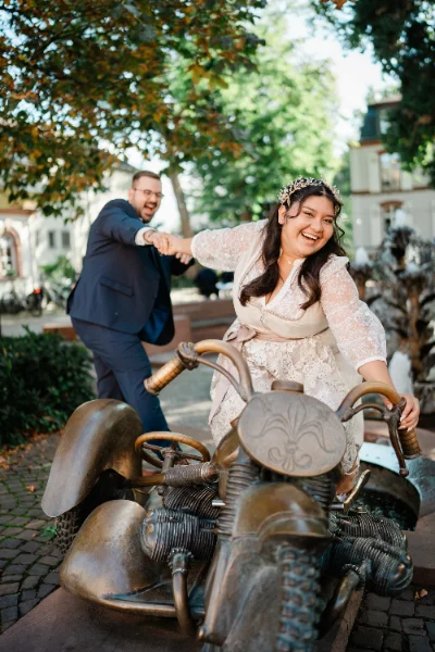 Brautpaar posiert lachend auf einer Motorrad-Skulptur aus Bronze